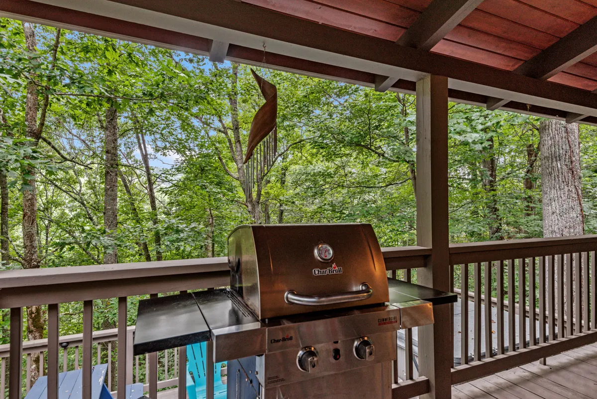 This image captures an outdoor grilling area on a wooden deck surrounded by lush greenery. The grill is prominently positioned near the railing, offering plenty of space for group meals and socializing. The casual setup invites guests to gather, share stories, and enjoy meals in the fresh mountain air.