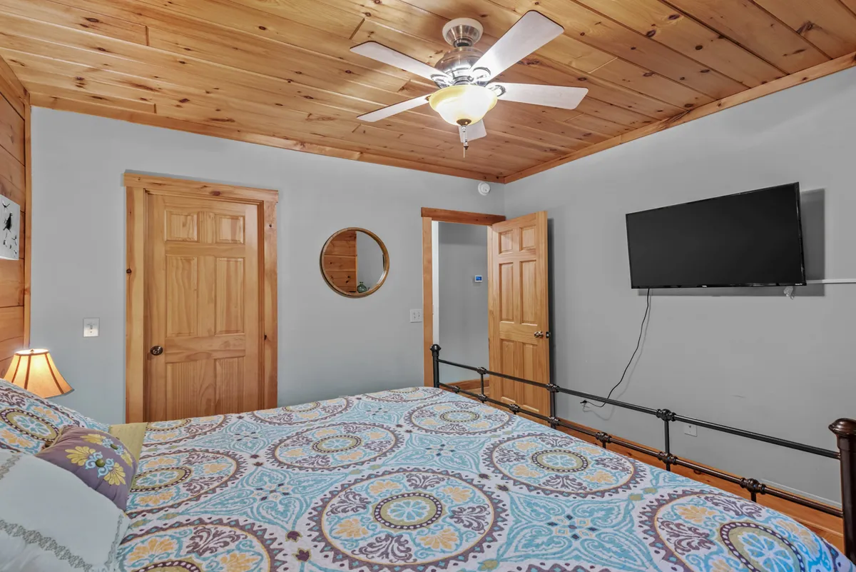A cozy bedroom with a king-size bed adorned with patterned bedding, wooden ceiling and walls, and a wall-mounted TV. A round mirror adds visual interest, and soft lighting creates a restful, inviting ambiance. The setting is perfect for relaxing, watching TV, or enjoying a quiet moment.