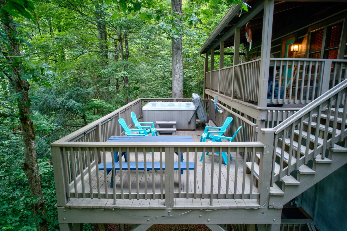 This exterior image features a spacious wooden deck equipped with a large hot tub and blue chairs, surrounded by lush greenery and forested views. The hot tub is prominently displayed, inviting guests to unwind and enjoy the natural setting in privacy.
