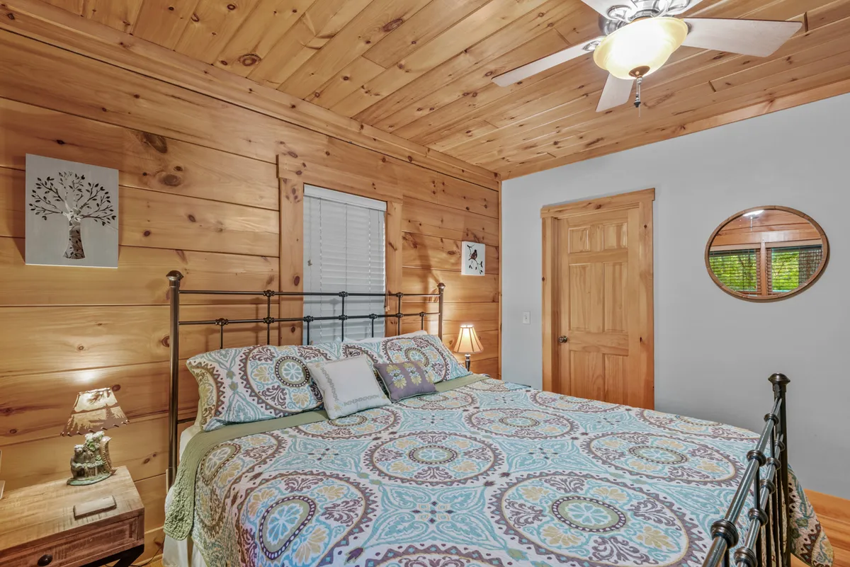 A cozy sleeping space with a comfortable bed dressed in colorful bedding, a nightstand with a lamp, and wood-paneled walls. The window offers a peek of sunlight, and the simple setup evokes warmth and flexibility for various sleeping arrangements.
