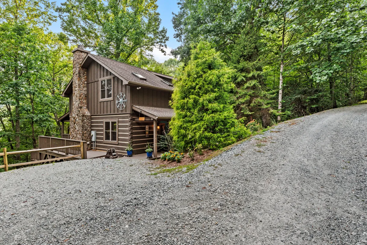 This image shows the cabin from an exterior perspective, framed by tall trees and a gravel path. The stone chimney, large porch, and surrounding natural landscape evoke a peaceful, mountain retreat ambiance.