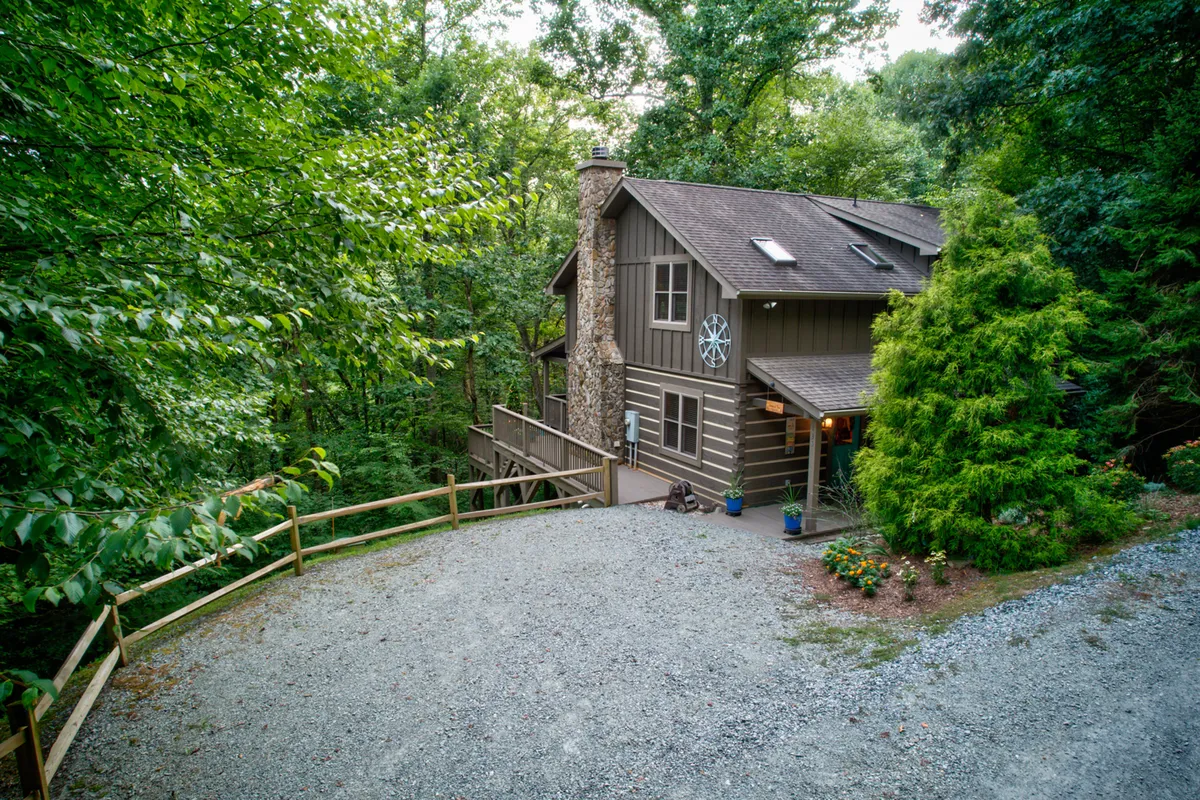 The image highlights the cabin's wooden exterior with a prominent stone chimney, set amid lush greenery and a gravel driveway. The peaceful setting hints at nature walks and the potential for outdoor activities nearby.