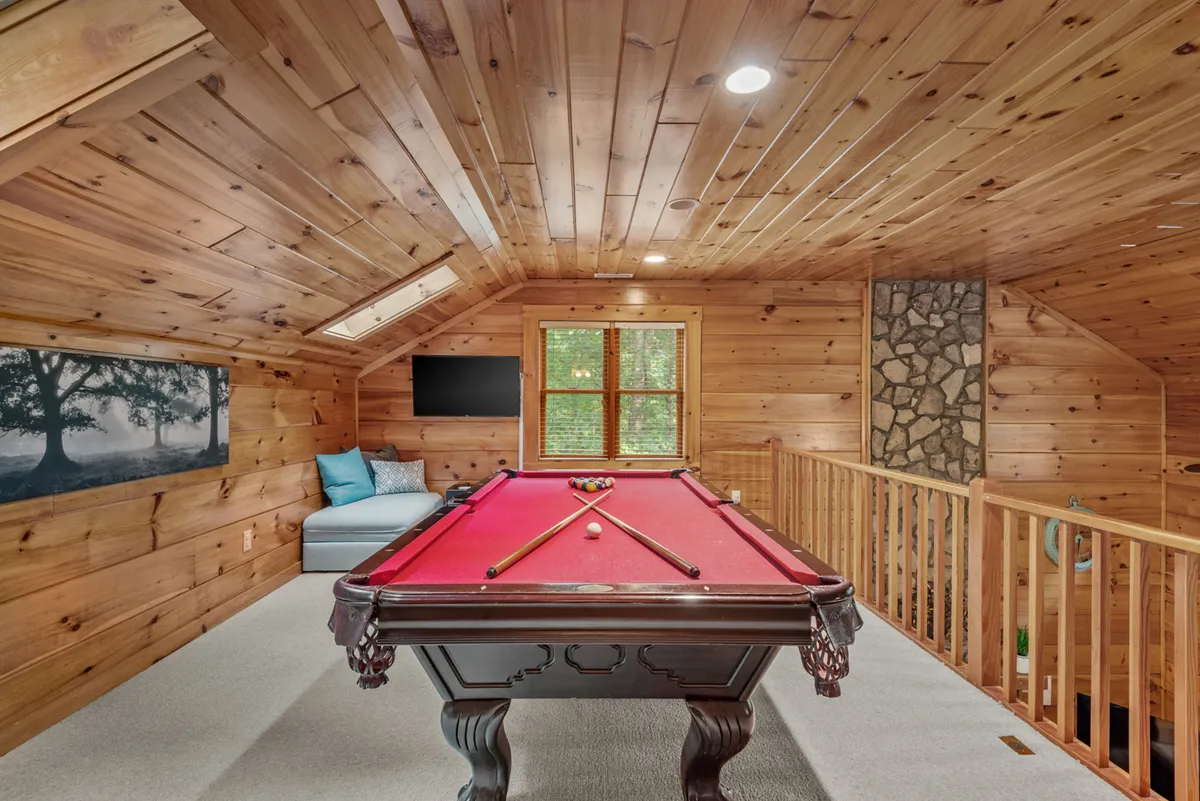 A spacious loft area with a full-size pool table sits beneath large windows that flood the space with natural light. Comfortable seating, including a sofa, is arranged nearby, with artwork on the walls adding a homey touch. The room is set up for both relaxation and friendly competition.
