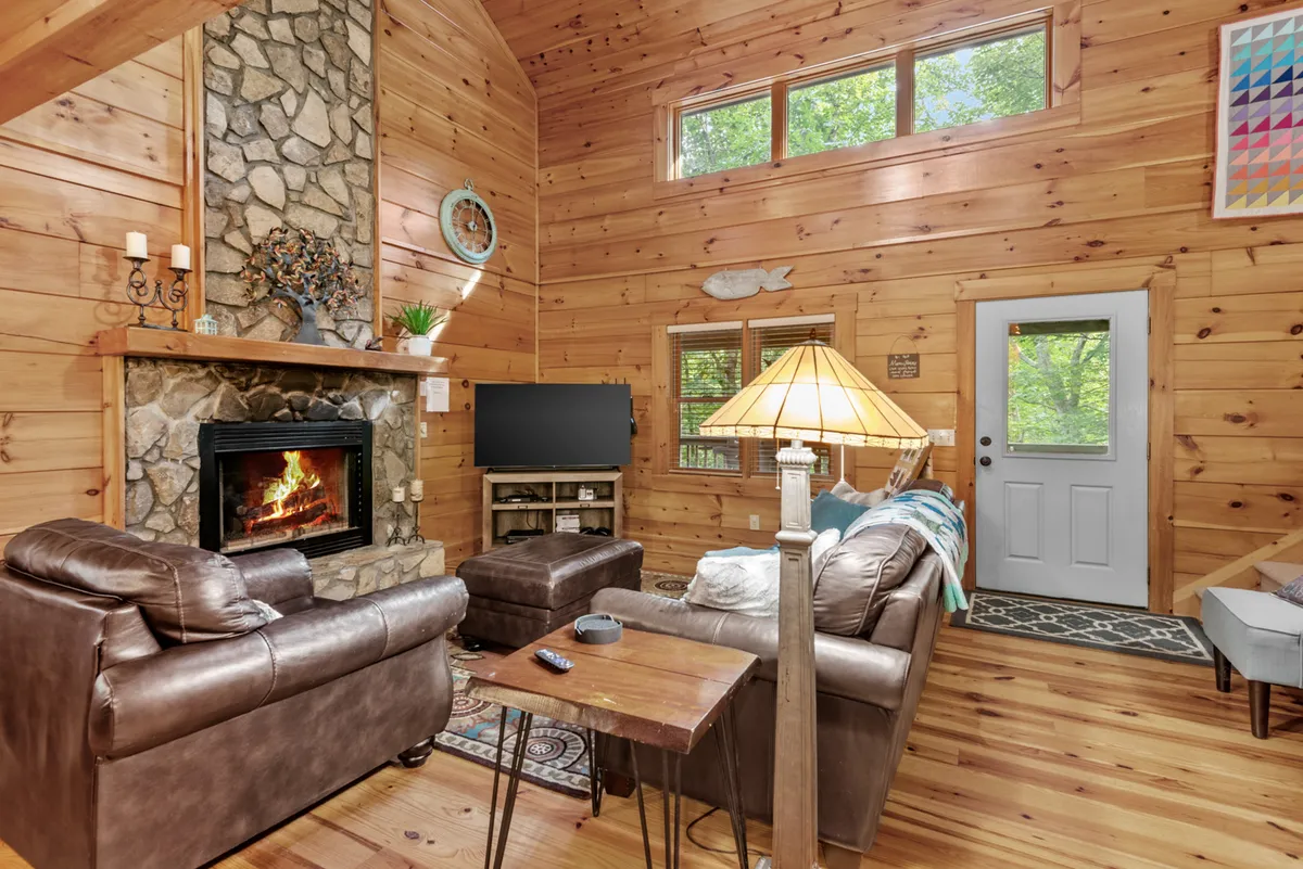 A welcoming living room features a striking stone gas-log fireplace as the central focal point, with plush leather sofas arranged for comfort. Large windows let in abundant natural light, highlighting the warm wood accents and providing a view of the surrounding landscape. A wooden table and decorative lamps add to the cozy, inviting atmosphere.