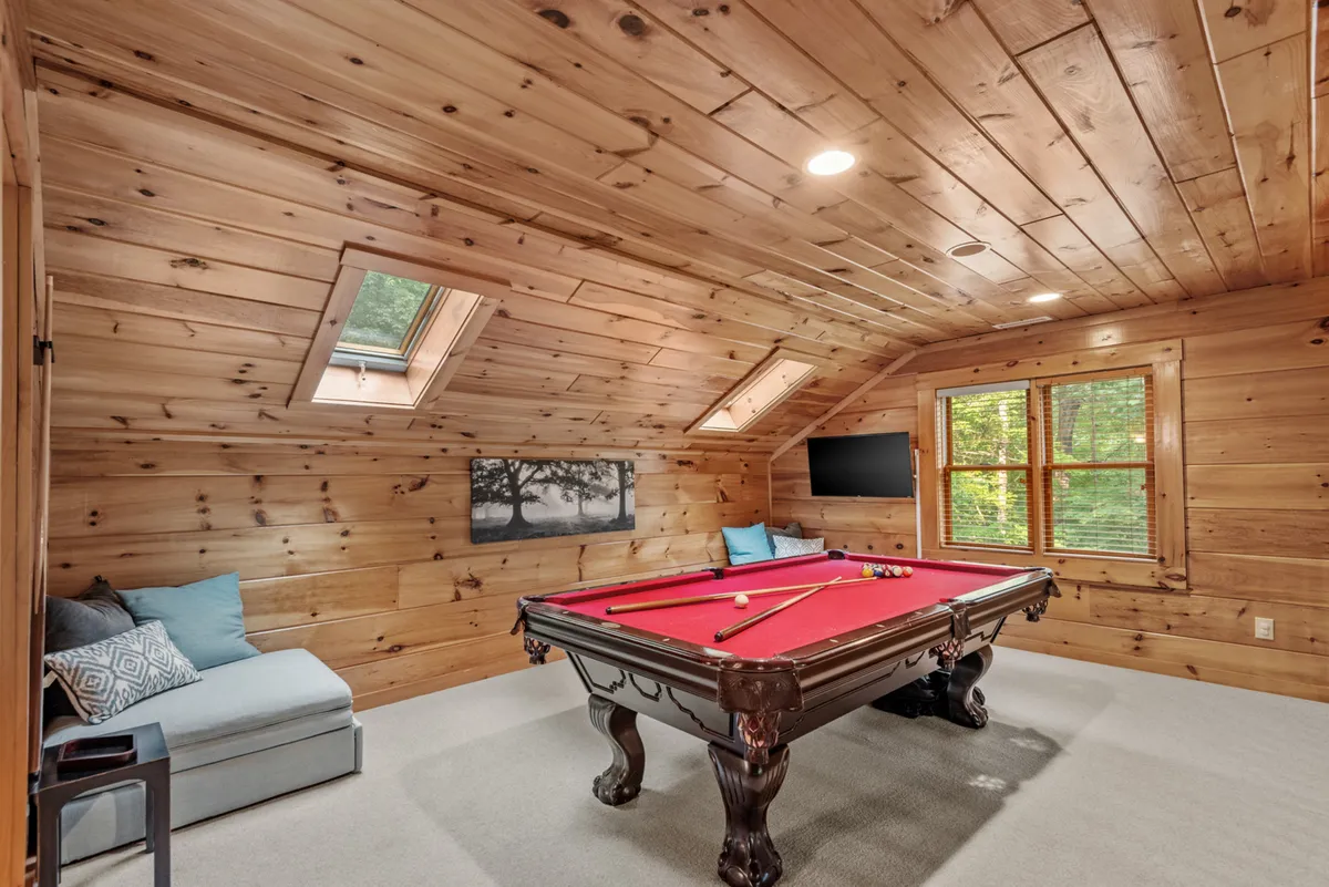 A bright, inviting game loft features a full-size pool table under skylights, with cozy seating and a television nearby. Wooden walls and comfortable furniture create a playful yet relaxed environment ideal for both spirited games and family gatherings.