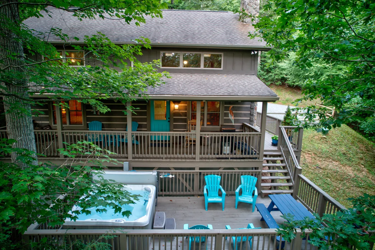 A tranquil exterior shot featuring a spacious wooden deck with a prominent hot tub surrounded by lush green foliage. Blue chairs and a sturdy deck railing invite relaxation and socializing, while the natural forest backdrop sets a peaceful mood.