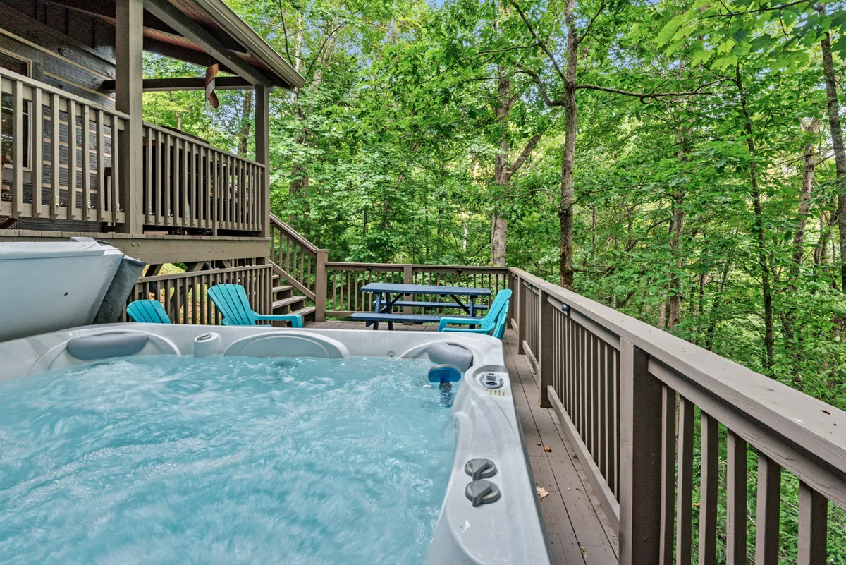 This photo features a spacious wooden deck with a hot tub, outdoor seating, and surrounding trees. It evokes the idea of relaxation, socializing, and enjoying nature together.