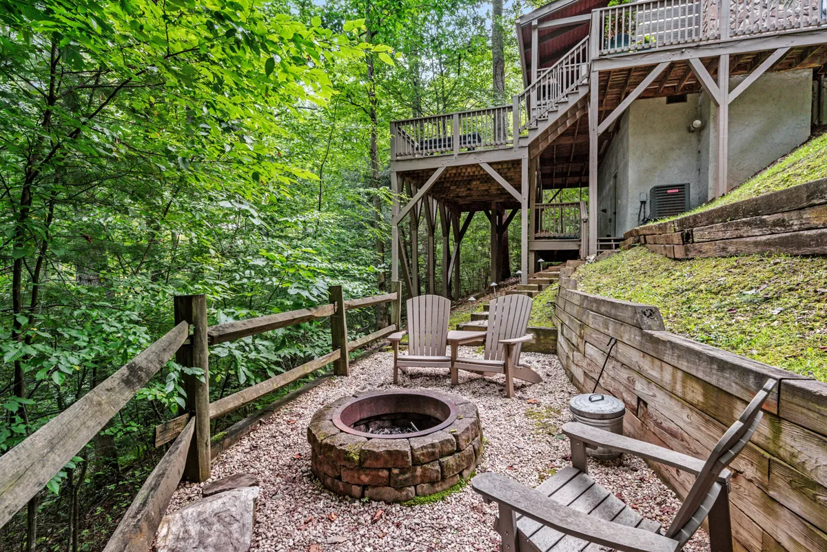 A cozy fire pit area with wooden chairs arranged for conversation, encircled by lush greenery and backed by the deck area. The scene is perfect for evening gatherings around the fire.