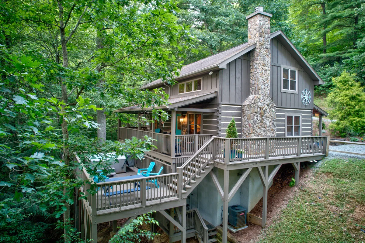 The image shows a charming wooden cabin nestled among lush green trees, featuring a spacious deck with inviting blue chairs, a striking stone chimney, and hints of a fire pit area. The setting emanates tranquility, with the deck positioned perfectly for enjoying sunset views and the surrounding natural beauty.