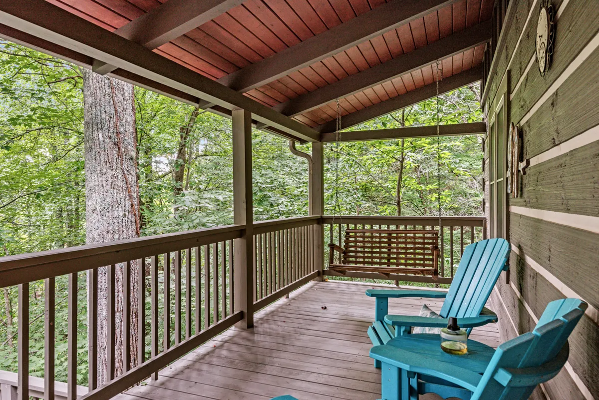 A cozy exterior porch scene featuring blue rocking chairs and a swing bench, all overlooking the surrounding trees. The setting suggests a peaceful spot for morning coffee, evening wine, or simply soaking up the quiet atmosphere of the woods.